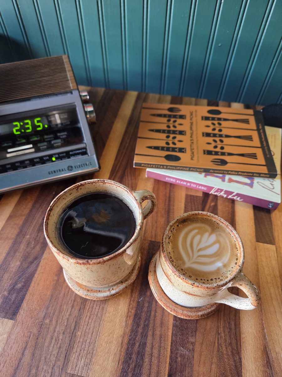 Drip coffee and a latte on a wooden table beside a stack of books